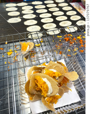 Thai street food featuring traditional coconut pancakes filled with shredded coconut and orange sweet topping on a market stall with cooking process visible Thai street food featuring traditional coconut pancakes filled with shredded coconut and orange sweet topping on a market stall with cooking process visible 137079767