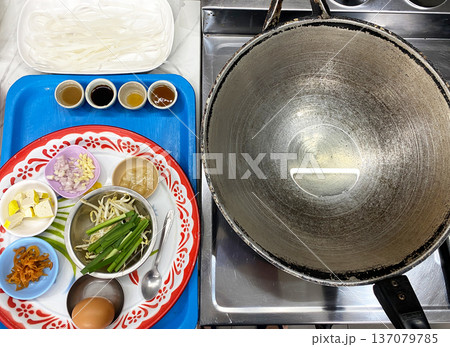Ingredients for making classic Pad Thai arranged on a decorative plate with a large wok and cooking utensils visible in a kitchen setting Ingredients for making classic Pad Thai arranged on a decorative plate with a large wok and cooking utensils visible in a kitchen setting 137079785