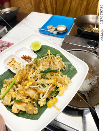 Plate of pad thai with chicken, bean sprouts, and lime served on banana leaf, with condiments and cooking equipment visible in the background 137079786