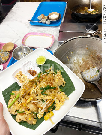 Plate of Pad Thai with chicken, bean sprouts, and lime served on banana leaf, cooking utensils and ingredients visible in a modern kitchen setting 137079787