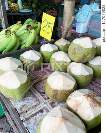 Fresh green coconuts with cut tops displayed on a market table, with a price sign and bunch of bananas in the background under natural lighting Fresh green coconuts with cut tops displayed on a market table, with a price sign and bunch of bananas in the background under natural lighting 137079822
