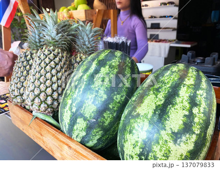 Freshly harvested watermelons and pineapples displayed in a wooden crate at a juice stand with a woman preparing drinks in the background 137079833