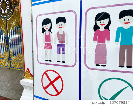 Warning sign at the entrance of a Thai temple displaying prohibited and permitted attire with illustrated figures and colorful graphics against a decorative background Warning sign at the entrance of a Thai temple displaying prohibited and permitted attire with illustrated figures and colorful graphics against a decorative background 137079834