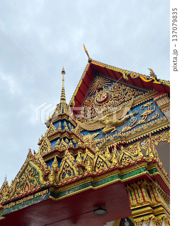 Ornate temple structure in Thailand featuring intricate gold decorations, colorful murals, and traditional architectural elements against a cloudy sky backdrop 137079835