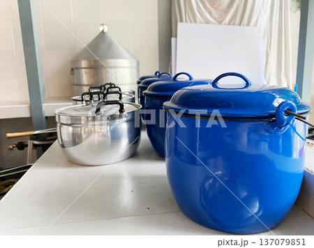 Blue and silver cooking pots arranged on a kitchen countertop with a large metal container and white paper backdrop visible in the background 137079851