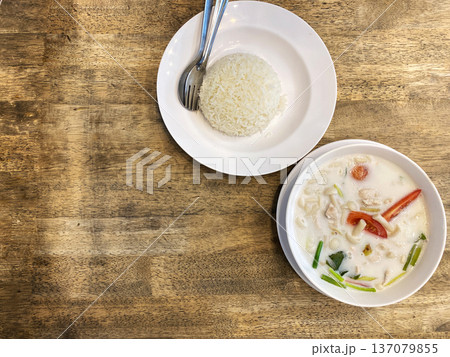 Coconut tom yum soup served in a bowl alongside a portion of steamed rice on a wooden table, showcasing vibrant ingredients and textures Coconut tom yum soup served in a bowl alongside a portion of steamed rice on a wooden table, showcasing vibrant ingredients and textures 137079855