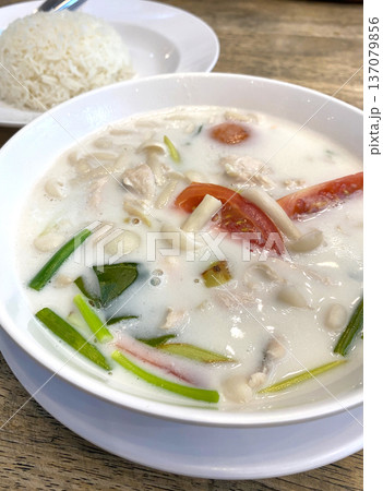 Bowl of creamy coconut soup with chicken, tomatoes, and herbs served alongside a portion of steamed rice on a wooden table 137079856