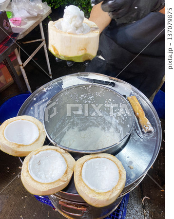 Coconut ice cream being served in a coconut shell, with multiple empty coconut shells on a metal container, showcasing a street food preparation scene 137079875