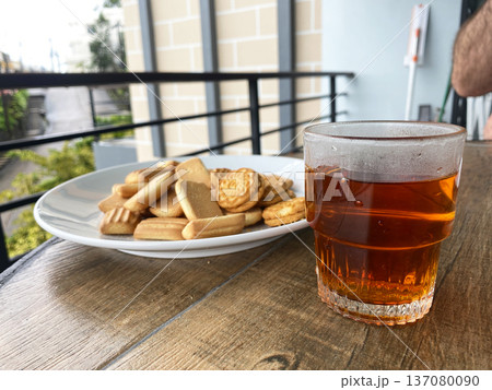 Glass of amber tea beside a plate of assorted cookies on a wooden table with a blurred outdoor background featuring greenery and a railing Glass of amber tea beside a plate of assorted cookies on a wooden table with a blurred outdoor background featuring greenery and a railing 137080090