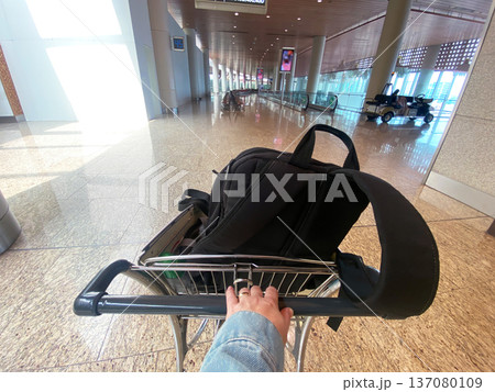 Woman's hand grips a luggage cart with a black backpack inside an airport terminal, bright lighting and modern architecture visible in the background Woman's hand grips a luggage cart with a black backpack inside an airport terminal, bright lighting and modern architecture visible in the background 137080109