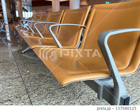 Modern airport seating arrangement featuring sleek orange leather chairs with metal armrests positioned on polished granite flooring in a spacious terminal area 137080125