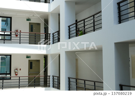Modern architectural interior of a multi-story building featuring open balconies, white walls, and metal railings with natural light illuminating the space Modern architectural interior of a multi-story building featuring open balconies, white walls, and metal railings with natural light illuminating the space 137080219