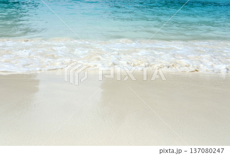 Calm turquoise ocean waves gently lapping onto a pristine sandy beach under bright sunlight, creating a serene coastal landscape with clear blue skies 137080247