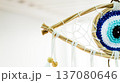 Crocheted evil eye dreamcatcher featuring a detailed blue and white eye pattern within a woven wooden frame and hanging white ribbons with wooden beads. Symbolizing protection and spiritual well being 137080646