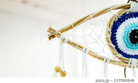 Crocheted evil eye dreamcatcher featuring a detailed blue and white eye pattern within a woven wooden frame and hanging white ribbons with wooden beads. Symbolizing protection and spiritual well being 137080646