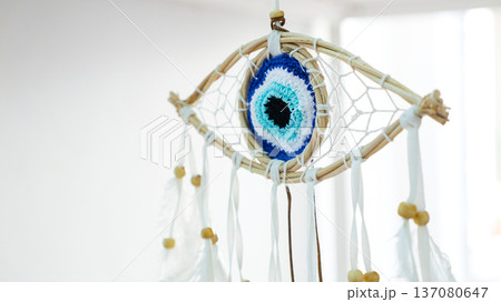 Handmade dreamcatcher amulet with an evil eye nazar woven from natural thread, accented with white feathers and wooden beads, hanging against a bright blue white backdrop 137080647