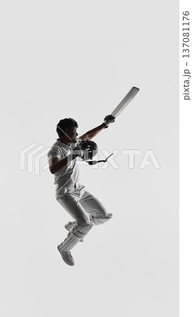 Cricket player jumping in air holding bat and helmet on white studio background in full white uniform. 137081176