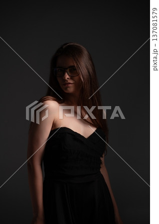 Stylish woman wearing glasses and black strapless dress posing with long brown hair, creating mysterious and elegant low key studio portrait for fashion and beauty concepts Stylish woman wearing glasses and black strapless dress posing with long brown hair, creating mysterious and elegant low key studio portrait for fashion and beauty concepts 137081579