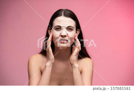Woman applying a refreshing sheet face mask for clear, glowing skin, focusing on beauty, self-care, and a healthy skincare routine against a pink background Woman applying a refreshing sheet face mask for clear, glowing skin, focusing on beauty, self-care, and a healthy skincare routine against a pink background 137081581
