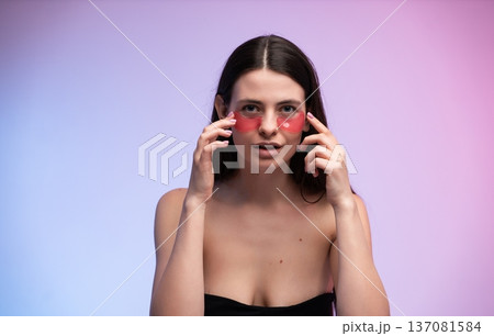 Young woman applying red hydrogel eye patches for under-eye skincare treatment, pampering and hydrating her skin against a gradient background Young woman applying red hydrogel eye patches for under-eye skincare treatment, pampering and hydrating her skin against a gradient background 137081584