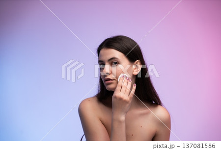 Woman cleansing her face with a cotton pad, performing daily skincare routine and makeup removal on a purple gradient background, illustrating beauty and hygiene Woman cleansing her face with a cotton pad, performing daily skincare routine and makeup removal on a purple gradient background, illustrating beauty and hygiene 137081586