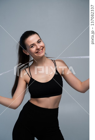 Young woman confidently holding a measuring tape draped around her neck, smiling brightly and showcasing fitness goals, body progress, and a healthy lifestyle Young woman confidently holding a measuring tape draped around her neck, smiling brightly and showcasing fitness goals, body progress, and a healthy lifestyle 137082337