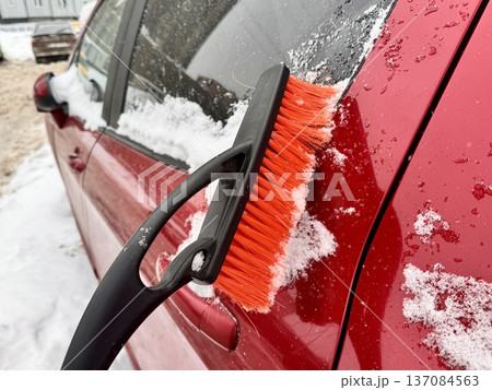 a girl cleans her personal car from snow with a black-and-orange car brush in the courtyard of an apartment building 137084563
