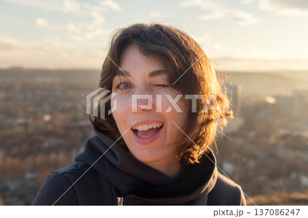 Young woman with short brown hair winks playfully at the camera, wearing a black hoodie, with a scenic city landscape and cloudy sky in the background 137086247