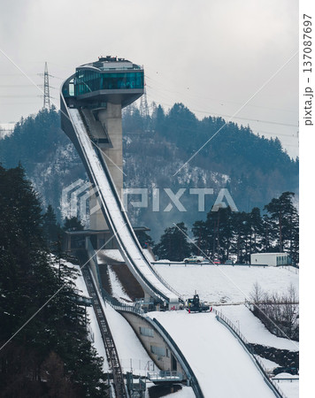 Aerial view of Bergisel Ski Jump in Innsbruck, Austria, with glass tower, sweeping ramp, snowy hills, power lines, groomed tracks, and machinery preparing the run. 137087697