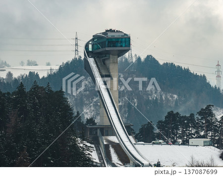 Ground level view of Bergisel Ski Jump in Innsbruck, Austria, steep inrun and glass deck visible, overcast sky, snow on pines, distant power lines and vehicles. Ground level view of Bergisel Ski Jump in Innsbruck, Austria, steep inrun and glass deck visible, overcast sky, snow on pines, distant power lines and vehicles. 137087699