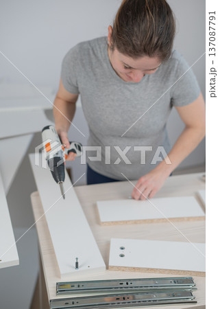 A Caucasian woman uses a screwdriver to tighten a screw while assembling a cabinet. A Caucasian woman uses a screwdriver to tighten a screw while assembling a cabinet. 137087791