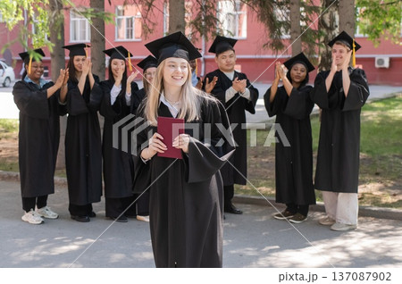 Young Caucasian woman holding diploma while classmates applaud her.  137087902