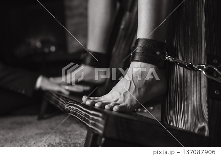 A man chains a woman's legs to a BDSM cross. A man chains a woman's legs to a BDSM cross. 137087906