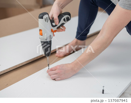 A Caucasian woman uses a screwdriver to tighten a screw while assembling a cabinet. A Caucasian woman uses a screwdriver to tighten a screw while assembling a cabinet. 137087941
