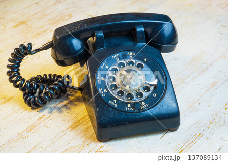 A black rotary phone with a coiled cord rests on a wooden table. The setting shows a simple room with natural light coming through a nearby window. 137089134