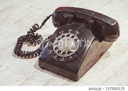 A rotary phone sits on a wooden table. The phone has a black body and a coiled cord. The background shows a light wooden surface that highlights the phone. 137089135