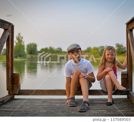 A little boy and girl sit on a wooden log on the shore of a lake. Small children sit on a pier at the edge of a pond. The concept of a carefree childhood and summer vacation. 137089503