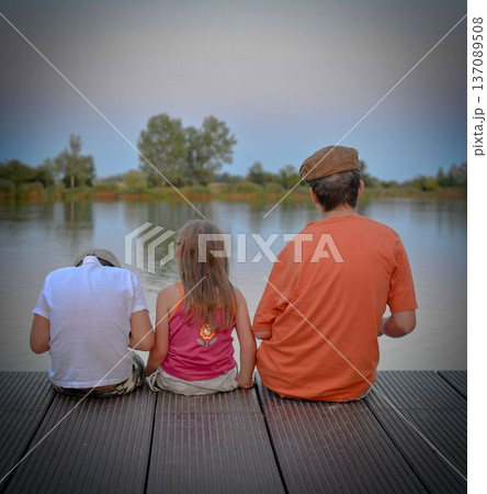 Children sitting on pier. Three children of different age - teenager boy, elementary age boy and preschool girl sitting on a wooden pier. Summer and childhood concept. Children on bench at the lake Children sitting on pier. Three children of different age - teenager boy, elementary age boy and preschool girl sitting on a wooden pier. Summer and childhood concept. Children on bench at the lake 137089508