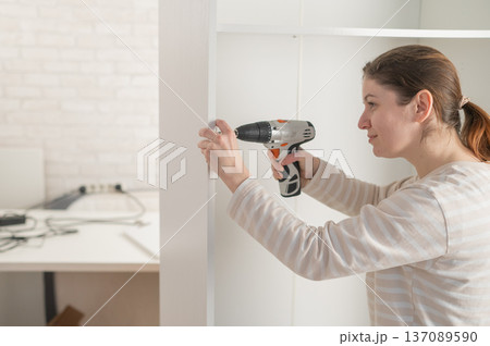 A Caucasian woman uses a screwdriver to tighten a screw while assembling a cabinet. A Caucasian woman uses a screwdriver to tighten a screw while assembling a cabinet. 137089590