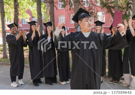 Young Asian man holds diploma while classmates applaud her.  137089648
