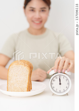 Happy smiling woman with whole wheat bread and alarm clock. concept of intermittent fasting, IF, meal timing, and choosing healthy carbohydrates for a balanced diet and weight loss management Happy smiling woman with whole wheat bread and alarm clock. concept of intermittent fasting, IF, meal timing, and choosing healthy carbohydrates for a balanced diet and weight loss management 137096118