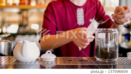 Tea shop. Preparing sachet drink. Barista pouring by spoon dried leaves in paper filter pack. Male worker measuring portion for herbal infuser. Tea shop. Preparing sachet drink. Barista pouring by spoon dried leaves in paper filter pack. Male worker measuring portion for herbal infuser. 137096609