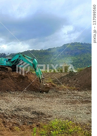 Green Excavator Working on Construction Site with Lush Forest Hill Background Green Excavator Working on Construction Site with Lush Forest Hill Background 137099560