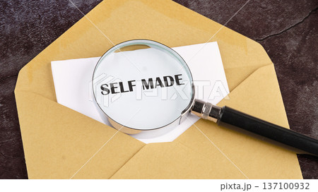 A bright magnifying glass reveals the profound words self made on a crisp white paper, nestled within a simple brown envelope on a textured dark background 137100932