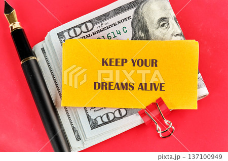 A vibrant red surface displays a stack of US hundred dollar bills, a sleek fountain pen, and a yellow card urging Keep Your Dreams Alive A vibrant red surface displays a stack of US hundred dollar bills, a sleek fountain pen, and a yellow card urging Keep Your Dreams Alive 137100949