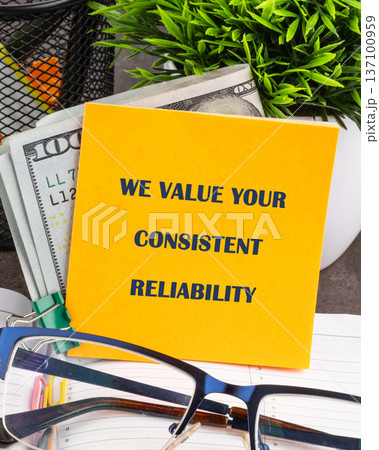 A vibrant yellow note praises consistent reliability. It rests with crisp US currency by a green plant, eyeglasses nearby on an open planner A vibrant yellow note praises consistent reliability. It rests with crisp US currency by a green plant, eyeglasses nearby on an open planner 137100959