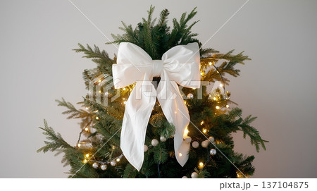 large white bow on the top of an evergreen tree, decorated with lights and garland, tied in the front with thick fabric 137104875