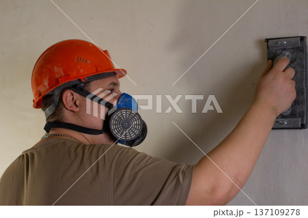 A construction worker wearing personal protective equipment, including a hard hat and a respirator, sands the plaster of an interior wall. Leveling the wall with a sanding machine. 137109278