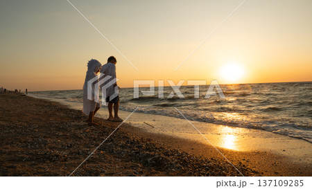 Two children, one wearing a hooded towel, standing by the water's edge on a pebble beach, watching the setting sun over the ocean waves during a summer vacation. Two children, one wearing a hooded towel, standing by the water's edge on a pebble beach, watching the setting sun over the ocean waves during a summer vacation. 137109285