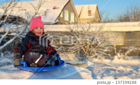 Cheerful young child wearing a pink hat and winter clothing, sitting on a blue sled, laughing and playing on a snowy outdoor day with houses and bare trees in background. 137109288